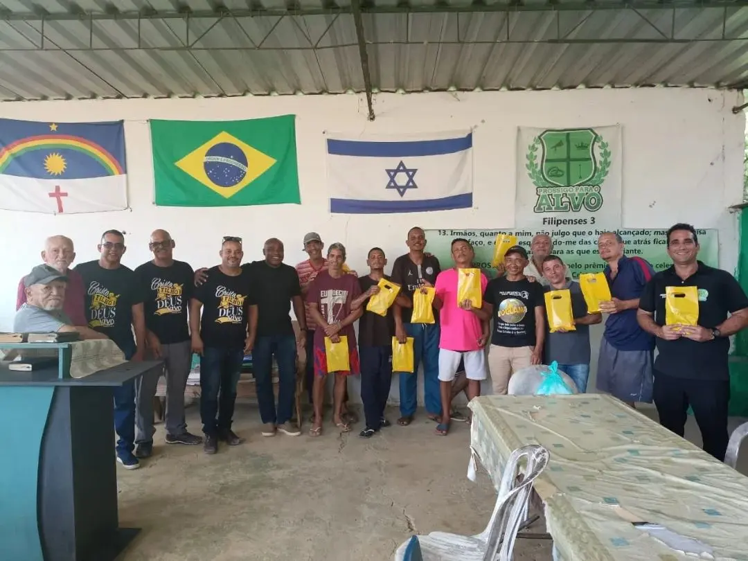 Fila de homens sorrindo, cada um segurando uma sacola amarela, durante uma ação da comunidade sob a faixa com o versículo de Filipenses 3.