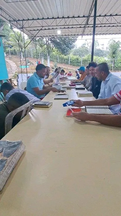 Homens sentados a uma longa mesa ao ar livre, lendo a Bíblia durante um estudo em grupo.